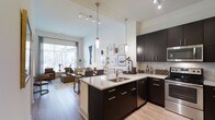 This stylish single-floor space offers a seamlessly integrated open-plan layout that combines the kitchen, dining, and living areas into one cohesive environment. The kitchen is a standout feature, showcasing rich dark cabinetry with sleek chrome hardware, light granite countertops, and a central island with a double sink — ideal for both meal preparation and casual dining. A marble-style tile backsplash adds a refined touch, while pendant lighting and recessed track lighting illuminate the space with both task and ambient light. The soaring ceilings amplify the sense of openness and airiness throughout. Wide-plank hardwood flooring runs continuously across the space, lending warmth and cohesion to the overall design. The living area benefits from generous natural light streaming through large sliding glass doors, offering views of the outdoors and creating a bright, welcoming atmosphere. A curated gallery wall adds a personal, artistic character to the dining and kitchen transition zone. The living area is generously proportioned, comfortably accommodating a full seating arrangement. Overall, the space strikes a balance between modern sophistication and everyday livability, with high-end finishes and thoughtful design details throughout.