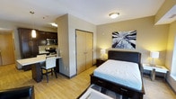 This single-floor studio is thoughtfully designed to maximize functionality within a compact footprint. The open-concept layout seamlessly integrates the sleeping, dining, and kitchen areas into one cohesive living space. Light hardwood-style flooring runs throughout, lending warmth and visual continuity across the entire unit.

The sleeping area is anchored by a dark-framed platform bed with built-in storage drawers, complemented by a matching nightstand and a softly glowing bedside lamp. A striking black-and-white nature-inspired artwork adorns the wall, adding a decorative focal point. A set of double doors provides access to a closet, offering practical storage solutions.

Adjacent to the sleeping area, a built-in desk with a bar-height chair serves as a dedicated workspace or dining nook. The kitchenette is efficiently outfitted with dark cabinetry, a stainless steel microwave, an electric stove, and a compact counter — providing essential cooking amenities in a well-organized arrangement.

Flush ceiling lighting ensures even illumination throughout the space. The overall ambiance is clean, modern, and functional, making it well-suited for individuals seeking a self-contained living environment.
