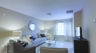 This single-floor space offers a warm and inviting atmosphere defined by a clean, contemporary aesthetic. The living area serves as the focal point of the space, showcasing soft, neutral wall tones in muted grays and whites that create a calm and sophisticated ambiance. Large windows dressed with sheer white curtains and horizontal blinds allow an abundance of natural light to filter through, brightening the room and enhancing its airy feel.

The living room is thoughtfully appointed with a plush light gray sofa accented by patterned throw pillows in navy and cream, complemented by additional seating that reinforces the room's cohesive color palette. A round white coffee table and a sleek glass-and-metal side table provide functional elegance, while a circular wall mirror with a metallic frame adds depth and visual interest to the space. A soft area rug anchors the seating arrangement, adding texture underfoot.

Recessed HVAC vents are integrated seamlessly into the ceiling, maintaining the clean lines of the interior. The overall design balances practicality with refined style, making this a comfortable and well-curated space suited for relaxed everyday living.