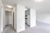 This single-floor unit offers a functional and straightforward layout designed to maximize usable living space. The living area is generously sized, finished with neutral-toned wall-to-wall carpeting that extends throughout the main spaces, and features a sliding glass door that opens to an outdoor patio area, allowing natural light to fill the room and providing views of surrounding greenery. A second window in what appears to be a bedroom also brings in outdoor light, overlooking the exterior surroundings.

The galley-style kitchen is positioned off the main living area, separated by a partial wall. It features gray cabinetry on both sides, dark laminate countertops, a white gas range, and a stainless steel sink. Light tile flooring distinguishes the kitchen from the carpeted areas of the unit.

The bedroom is a well-proportioned room with soft carpeting and a reach-in closet, offering practical storage. The bathroom includes a full tub and shower combination with white tile surround, a toilet, vanity with sink, and a distinctive mosaic tile floor in a blue and white pattern, adding a subtle decorative element to the otherwise neutral palette throughout the unit.