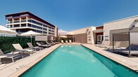 This outdoor pool area offers a resort-style retreat designed for relaxation and recreation. At its center sits a generously sized rectangular lap pool with crystal-clear turquoise water, surrounded by a wide, light-colored concrete deck that provides ample space for lounging and movement. Numerous adjustable chaise lounges are arranged along all sides of the pool, offering guests a variety of spots to unwind in the sun. Several large market umbrellas provide shaded seating areas throughout the deck, while a pair of sleek cabana-style daybeds offer a more private lounging experience. A lush green privacy hedge encloses portions of the perimeter, creating a sense of seclusion while still allowing for sweeping views of the surrounding mountains — visible in the background with snow-capped peaks on clear days. The pool deck is serviced by a modern single-story amenity building featuring glass double-entry doors, providing convenient access to interior facilities. The overall aesthetic is clean and contemporary, with a neutral palette of tans, whites, and grays that complement the natural landscape. The combination of open sky, mountain vistas, and thoughtfully arranged amenities gives this pool deck a bright, airy, and inviting ambiance suited for both leisure and active use.