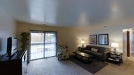 This single-floor space offers a thoughtfully arranged and comfortable living environment. The living area serves as the centerpiece of the space, featuring soft wall-to-wall carpeting that adds warmth underfoot. A large sliding glass door floods the room with natural light and provides direct access to an outdoor balcony, offering views of the surrounding greenery. Vertical blinds on the sliding door allow for easy light control and privacy. The room is finished with a textured ceiling that adds subtle architectural character. Furnishings include a plush, dark gray sofa paired with a patterned accent chair, a sleek glass-top coffee table, and a coordinating area rug that anchors the seating arrangement. A pair of framed wall art pieces and a classic table lamp with warm ambient lighting complete the refined, cozy aesthetic. The neutral, warm-toned walls provide a versatile backdrop that complements a variety of décor styles. Overall, the space strikes a balance between functionality and comfort, making it an inviting and practical living environment with a welcoming ambiance.