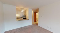 This single-floor unit offers a functional, open layout designed for comfortable everyday living. The living area is generously proportioned, finished with neutral-toned carpet throughout, and anchored by a sliding glass door that opens to a private balcony, flooding the space with natural light and providing outdoor views.

The kitchen is positioned adjacent to the living area and is accessible via a pass-through counter with a raised bar ledge, creating a connected yet defined cooking space. White cabinetry lines the walls, accompanied by a double stainless steel sink, a gas range, and a refrigerator. A ceiling fan with a light fixture overhead provides both ventilation and ambient lighting. Tile flooring runs throughout the kitchen, offering a practical contrast to the carpeted living spaces.

A dedicated bedroom is accessible via a hallway and features its own window with horizontal blinds, allowing natural light to filter in while maintaining privacy. The neutral white walls and carpet create a calm, understated atmosphere.

The bathroom includes a single vanity with cabinet storage, a medicine cabinet mirror, a toilet, and a tiled shower-tub combination, all finished in a clean white palette with classic tile flooring.