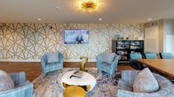 This club lounge is a thoughtfully designed social space that blends contemporary style with comfortable functionality. The centerpiece of the room is a striking accent wall adorned with an angular gold geometric pattern, providing a bold visual backdrop for the large wall-mounted flat-screen television. A statement sputnik-style gold ceiling fixture casts warm light throughout the space, complementing the rich hardwood-style flooring underfoot.

The lounge area is furnished with plush, blue-grey upholstered chairs arranged around a sculptural white coffee table and paired with sleek gold side tables, creating an inviting setting for relaxed gathering. A dining area with a rustic wood table and additional seating offers a more structured space for group use. Along one wall, a snack and beverage station provides convenient refreshments, while a built-in kitchenette with cabinetry and appliances adds practical utility.

Large sliding glass doors flood the interior with natural light and open directly onto a furnished outdoor terrace. The terrace features comfortable seating arrangements and open skyline views, extending the usable social space outdoors. Together, the interior and exterior areas create a versatile, upscale environment well-suited for both casual lounging and organized gatherings.