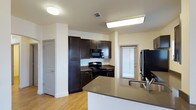This single-floor space centers around a thoughtfully designed kitchen that serves as its standout feature. The kitchen is equipped with dark espresso-toned cabinetry that extends from floor to ceiling, offering generous storage capacity. Black appliances, including an electric range, over-the-range microwave, and refrigerator, complement the dark cabinet palette and lend a cohesive, modern aesthetic to the space.

A peninsula with a smooth gray countertop extends into the open area, providing additional workspace as well as a sink with a contemporary brushed-metal faucet. This layout supports an open-concept flow, making the kitchen both practical and accessible from multiple angles.

The flooring throughout is light wood-tone laminate or hardwood, which brightens the interior and contrasts effectively with the darker cabinetry. Walls are finished in a clean, neutral off-white tone. Overhead, flush-mount and recessed fluorescent fixtures provide ample artificial lighting, while a window with horizontal blinds near the cooking area admits natural light. The overall ambiance is clean, functional, and contemporary.