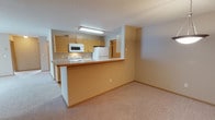 This single-floor residence offers a practical and functional layout centered around an open-concept design that seamlessly connects the kitchen and living areas. The kitchen is equipped with light oak cabinetry providing ample storage, white appliances including a range, microwave, and refrigerator, and is illuminated by a flush fluorescent light fixture. A peninsula-style breakfast bar extends from the kitchen, creating a natural division between the cooking space and the carpeted living area while maintaining an open, airy feel. The warm-toned countertop on the breakfast bar adds a subtle contrast to the neutral wall color, which carries throughout the space in soft beige and gray hues. The living area features wall-to-wall carpet in a light neutral tone, contributing to a cozy and understated ambiance. A hallway visible beyond the kitchen leads to additional areas of the residence, suggesting a thoughtful floor plan that balances open communal spaces with more private quarters. The textured ceiling and clean lines of the walls lend a simple, unpretentious character to the overall interior. This space is well-suited for those seeking a straightforward, move-in ready layout with cohesive finishes and an efficient use of space.