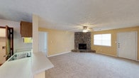 This single-floor home offers a comfortable and functional layout designed for easy living. The main living area is a standout space, characterized by warm yellow walls, plush wall-to-wall carpeting, and a textured ceiling that adds a classic touch. A striking natural stone corner fireplace serves as the focal point of the room, providing both visual interest and a cozy atmosphere. A ceiling fan overhead ensures year-round comfort, while a large window allows natural light to fill the space. The open layout connects seamlessly to an adjacent area, visible through a doorway, suggesting a flexible floor plan that accommodates a variety of living arrangements. A partial counter is visible along one side, hinting at a nearby kitchen or bar area that integrates conveniently with the main living space. White baseboards and trim provide clean, crisp contrast against the warm-toned walls. Overall, the home presents a bright, inviting ambiance with a blend of rustic and traditional design elements that create a lived-in, welcoming feel throughout.