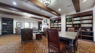 This elegant single-floor community space blends rustic charm with refined comfort through a cohesive Spanish Colonial design aesthetic. Exposed dark wood ceiling beams run throughout, complemented by rich hardwood built-in bookshelves, dark-framed French doors, and a distinctive bold floral-patterned tile floor in deep reds and golds that anchors the entire space.

The library and lounge area serves as the heart of the space, featuring a white fireplace with decorative tilework surround, built-in shelving stocked with books, and plush patterned armchairs arranged for intimate conversation. Natural light filters in generously through multiple sets of French doors, offering views to an outdoor balcony area. Geometric lantern-style pendant lights add warmth and character overhead.

A dedicated game area provides a change of pace, furnished with a chess table, cushioned barrel chairs, a large flat-screen television, and a granite-topped media console with ample drawer storage. The dollhouse view also reveals a full billiards table, adding to the recreational offerings.

A well-appointed wet bar area includes extended granite countertops, a stainless steel sink, dark wood cabinetry, wainscoting, and a refrigerator — ideal for hosting gatherings. Throughout, equestrian-themed artwork and lush greenery add curated character, creating a warm, club-like ambiance suited for relaxation and socializing alike.