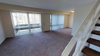This two-floor residential space offers a functional and comfortable layout designed to maximize natural light and open living. The main living area is a standout feature, showcasing wall-to-wall carpeting in a neutral tone that adds warmth underfoot. Large sliding glass doors and full-height vertical blinds line the exterior wall, allowing an abundance of natural light to flood the room while providing direct access to an outdoor area. The generous proportions of the living space create an open, airy feel with room for a variety of furniture arrangements. A central entry door is flanked by the expansive glazed panels, reinforcing the connection between interior and exterior. The soft, neutral wall tones throughout complement the carpeted floors and contribute to a calm, understated ambiance. A staircase with a clean white railing is visible, providing access between the two floors and adding a subtle architectural element to the space. Flush-mount ceiling lighting provides even illumination throughout the main level. Overall, the space presents a straightforward, well-lit residential environment with practical finishes and a layout that emphasizes openness and livability across both floors.