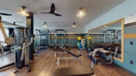 Located in Kingston, MA, this single-floor fitness center is designed to accommodate a wide range of workout needs in a thoughtfully appointed space. The interior is distinguished by a bold teal and gold horizontal stripe accent wall, warm wood-look flooring, and multiple ceiling fans with pendant-style lighting that create a comfortable and energized atmosphere.

The strength training area is well-stocked with a power rack, adjustable weight benches, a full dumbbell rack, cable cross machine, and a dedicated functional training rig. A large wall-mounted mirror enhances the space, allowing users to monitor form during lifts. Medicine balls and stability balls are neatly organized on a dedicated rack nearby.

The cardio section lines the windowed wall, where large windows allow abundant natural light to fill the room. Equipment includes treadmills, a rowing machine, and a cable/multi-functional machine, all positioned to take advantage of the bright, open feel of this side of the facility.

A built-in amenity station features a granite-topped counter with wood cabinetry, integrated storage lockers, a beverage refrigerator, and open metal shelving above stocked with towels and accessories. This practical station ensures members have everything they need within easy reach throughout their workout.