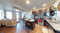 This single-floor space showcases an open-concept layout that seamlessly integrates the kitchen, dining, and living areas into one cohesive and functional environment. The kitchen is a standout feature, outfitted with stainless steel appliances — including a refrigerator, gas range, and built-in microwave — and rich dark wood cabinetry with ample storage. A central kitchen island with light countertops provides additional prep space and doubles as a casual dining area, complete with stylish bar-height seating. Warm hardwood flooring extends throughout the space, tying the rooms together with a consistent, inviting aesthetic. Exposed ductwork along the ceiling adds an industrial character that complements the modern finishes. The living area is bright and generously proportioned, benefiting from large windows that usher in natural light and offer views to the exterior. A ceiling fan contributes both function and style to the room. The overall ambiance is contemporary and well-appointed, striking a balance between urban industrial design elements and comfortable, practical living — making it an ideal space for both everyday living and entertaining.