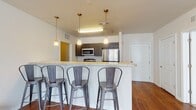 This single-floor unit is designed with an open-concept layout that seamlessly connects the kitchen, dining, and living areas. The kitchen is a functional centerpiece, equipped with stainless steel appliances including a refrigerator, microwave, and range. Dark espresso cabinetry contrasts with light granite countertops, and a spacious breakfast bar with seating for multiple guests serves as both a dining and gathering space. Pendant lighting suspended from above adds a warm, focused glow over the bar area.

The living room features rich hardwood-style flooring and benefits from natural light through a window overlooking the exterior. A ceiling fan provides additional comfort and airflow throughout the shared living space.

The bedrooms are carpeted for comfort and each includes a ceiling fan and window for natural light. One bedroom connects directly to a private bathroom, providing an en-suite arrangement. Both bathrooms are well-appointed with dark wood vanities, granite countertops, and a shower-tub combination. Wood-look tile flooring in the bathrooms complements the overall design aesthetic. Closet storage is available within the unit, contributing to the practical and cohesive character of this well-organized space.