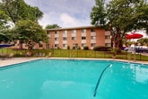 Located at 38 N Summit Dr in Gaithersburg, Maryland, this outdoor amenity space centers around a generously sized swimming pool with clear turquoise water. The pool is accessed via stainless steel entry rails and is enclosed by a black iron security fence, ensuring a safe and well-defined recreational environment. Surrounding the pool is a broad concrete sundeck that provides ample room for lounging, furnished with multiple reclining chairs and seating arrangements positioned to take advantage of the open-air setting. Patio umbrellas offer shaded spots throughout the deck area for added comfort. A dedicated support structure on one end of the pool area houses restroom and utility facilities, complementing the overall functionality of the space. Mature deciduous trees frame the perimeter, creating a lush, green canopy that adds privacy and a natural ambiance to the outdoor setting. In the background, a classic multi-story brick residential building with a uniform grid of windows overlooks the pool area, reinforcing the community-oriented character of the property. The combination of open sightlines, mature landscaping, and well-maintained facilities makes this outdoor pool area a practical and inviting communal space for residents of the Gaithersburg community.