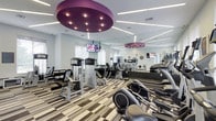 This contemporary fitness center offers a well-appointed, single-floor workout space designed to accommodate a full range of exercise needs. The room is anchored by bold, circular purple pendant light fixtures that hang from a white ceiling interlaced with linear LED strip lighting, creating a vibrant and energizing atmosphere. Large mirrors line the walls, visually expanding the space and allowing members to monitor their form throughout their workouts. The flooring features a stylish striped carpet with neutral tones, adding a modern touch underfoot. The facility is outfitted with a comprehensive selection of SportsArt brand equipment, including cable machines, strength-training stations, treadmills, ellipticals, and other cardio machines. Wall-mounted flat-screen televisions are positioned throughout the space, providing entertainment during workouts. Natural light filters in through windows, complementing the bright, well-lit interior. Circular decorative wall accents add an additional design element that ties the room's aesthetic together. Overall, the fitness center combines functionality with a polished, upscale design, making it a well-equipped destination for a complete workout experience.