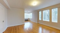 This single-floor space presents a clean, open layout characterized by warm hardwood flooring that runs throughout, reflecting natural light and adding a timeless quality to the interior. The main living area is generously proportioned, with crisp white walls and white baseboard trim that create a bright, neutral palette well-suited for a variety of uses and furnishing styles.

A standout architectural feature is the bay window area, which floods the space with abundant natural light from multiple angles while offering views of the surrounding greenery. An elegant pendant light fixture hangs within this alcove, adding a subtle decorative touch. Additional large windows along the adjacent wall further enhance the brightness and airiness of the room, ensuring consistent daylight throughout the day.

The textured ceiling adds a classic residential character, while the flush-mount ceiling fixture provides practical overhead lighting. A traditional cast-iron radiator is positioned beneath the bay window, serving as both a functional heating element and a nod to the space's classic architectural roots. The overall ambiance is light, open, and inviting, with a straightforward layout that maximizes usable floor space and lends itself well to flexible arrangements.
