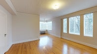 This single-floor space presents a clean, open layout characterized by warm hardwood flooring that runs throughout, reflecting natural light and adding a timeless quality to the interior. The main living area is generously proportioned, with crisp white walls and white baseboard trim that create a bright, neutral palette well-suited for a variety of uses and furnishing styles.

A standout architectural feature is the bay window area, which floods the space with abundant natural light from multiple angles while offering views of the surrounding greenery. An elegant pendant light fixture hangs within this alcove, adding a subtle decorative touch. Additional large windows along the adjacent wall further enhance the brightness and airiness of the room, ensuring consistent daylight throughout the day.

The textured ceiling adds a classic residential character, while the flush-mount ceiling fixture provides practical overhead lighting. A traditional cast-iron radiator is positioned beneath the bay window, serving as both a functional heating element and a nod to the space's classic architectural roots. The overall ambiance is light, open, and inviting, with a straightforward layout that maximizes usable floor space and lends itself well to flexible arrangements.