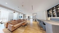 This thoughtfully designed single-floor amenity space offers a generous and open layout encompassing a living room, dining room, and fully equipped kitchen. The interior is defined by a clean, modern aesthetic, with light hardwood flooring flowing seamlessly throughout the main living and dining areas, complemented by crisp white walls and recessed lighting.

The expansive living room is anchored by a plush sectional sofa, a wooden coffee table, and stylish pendant lighting. Decorative lattice-style room dividers subtly zone the space while maintaining an open feel, and a wall-mounted TV adds a functional focal point. A built-in display shelf with accent lighting and curated artwork lend the space a gallery-like quality.

The dining area is a standout, featuring floor-to-ceiling windows dressed in sheer white curtains that flood the room with natural light. A large communal table paired with mixed seating and sculptural pendant fixtures creates an inviting setting for gatherings. A navy accent wall with framed artwork adds depth and visual interest.

The kitchen is efficiently laid out with light wood cabinetry, stainless steel appliances including a full-sized refrigerator, oven, and microwave, and neutral tile flooring. A separate bar counter with high stools provides an additional casual dining option. Overall, the space exudes a bright, contemporary, and welcoming ambiance.