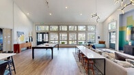 Located at 3280 SW 170th Ave in Beaverton, Oregon, this thoughtfully designed single-floor community space offers a generous and versatile layout spanning a substantial footprint. The open-concept floor plan seamlessly connects the kitchen and living areas, creating a cohesive and inviting environment.

The kitchen area is anchored by a large central island with a white quartz surface, surrounded by stylish dark wood bar seating. Rustic wood-toned cabinetry, white subway tile backsplash, and bold pendant lighting lend the space a contemporary yet warm character. Interior pass-through windows connect to adjacent rooms, enhancing the sense of openness.

The living area is a true showpiece, featuring soaring vaulted ceilings that amplify the sense of space. Floor-to-ceiling windows along an entire wall flood the room with natural light and offer views of the surrounding outdoor areas. A striking geometric color-block accent wall frames a large flat-screen display, complemented by sculptural branch-style chandeliers overhead. Comfortable sectional seating, plush area rugs, and curated artwork contribute to a vibrant, modern aesthetic.

A dedicated lounge nook with bold mural walls and booth-style seating provides an intimate gathering spot, while the wood-plank flooring and carpeted zones define each area with purposeful design throughout.