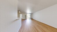 This single-floor unit offers a clean, modern aesthetic defined by light wood flooring that runs consistently throughout the space, crisp white walls, and an abundance of natural light. The open living area is generously proportioned, with a ceiling fan overhead and a sliding glass door that leads directly to an outdoor patio, allowing natural light to fill the room. A wall-mounted heating unit and a through-wall air conditioning unit ensure year-round comfort. A half-wall partition separates the living space from the galley-style kitchen, which features white shaker cabinetry with brushed hardware, light countertops, a gas range, a dishwasher, and a stainless steel sink — all finished with the same warm wood flooring for a cohesive look. The bedroom is a calm, well-lit retreat with multiple windows that frame outdoor greenery, a wide sliding-door closet providing ample storage, and a wall-mounted heating unit. The bathroom is clean and functional, featuring a full tub-shower combination with built-in shelving, all finished in white. Throughout the unit, a neutral palette and consistent materials create a bright, cohesive, and inviting atmosphere well-suited for comfortable everyday living.