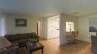This single-floor space offers an open and functional layout that connects the living area, kitchen, and a casual dining space. The living room is comfortably sized, featuring light hardwood flooring, diagonal wood-paneled walls, and a textured ceiling. A dark wood coffee table and upholstered sofa anchor the space, complemented by framed wall art that adds a personal touch. Natural and ambient lighting keeps the room feeling bright and welcoming.

A wide open passageway leads directly into the kitchen, which is outfitted with white cabinetry extending to the ceiling for ample storage. The countertops are clean and minimal, with a sink positioned beneath upper cabinets. The kitchen maintains the same light hardwood flooring, creating a cohesive flow throughout the space.

Adjacent to the kitchen, a pass-through window opens to a small breakfast bar area with bar stools, ideal for casual dining or entertaining. A potted plant on the bar ledge adds a touch of greenery. A hallway visible from the living room suggests additional rooms beyond, contributing to the overall practical and straightforward layout of this single-level space.