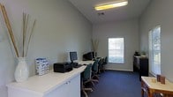 This single-floor space is designed as a practical and efficient computer lab or business center. The room features vaulted ceilings that lend a sense of openness despite the compact footprint, with neutral gray walls providing a clean and professional backdrop throughout. A long, white built-in desk runs along the perimeter of the room, offering generous workspace across multiple computer workstations. Each station is equipped with a desktop monitor and is paired with an ergonomic rolling chair upholstered in teal green, creating a cohesive and organized look. A printer and stacked supply boxes are positioned at one end of the desk, indicating the space is fully equipped for general office and business tasks. A window fitted with horizontal blinds allows natural light to filter into the room while maintaining privacy, complementing the overhead fluorescent lighting that ensures consistent illumination throughout. Blue-toned carpeting covers the floor, adding a layer of comfort underfoot. Decorative bamboo stalks in the corner provide a subtle touch of warmth to the otherwise utilitarian setting. Overall, the space is thoughtfully arranged to maximize productivity and functionality in a clean, well-lit environment.