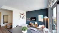 This well-appointed single-floor residence showcases a cohesive modern design throughout. The living area is anchored by a striking deep teal accent wall, which contrasts sharply against bright white walls and light wood-style flooring. Floor-to-ceiling windows flood the space with natural light while offering views of the surrounding urban landscape. Thoughtful design details, including wall-mounted bike storage and transit-themed artwork, give the living area a distinctly urban character.

The kitchen features a streamlined layout with high-gloss white upper cabinetry paired with dark wood-toned lower cabinets. A granite backsplash and matching countertops add texture and refinement, while stainless steel appliances, including a gas cooktop and built-in oven, provide a fully functional cooking space.

The bedroom continues the teal accent wall motif, creating a consistent design language across the unit. A large floor mirror and recessed lighting contribute to a bright, well-proportioned feel.

The bathroom maintains the modern aesthetic with neutral tile work, a dark-toned vanity with a white countertop, a soaking tub with an integrated shower, and in-unit stacked laundry conveniently accessible nearby.