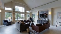 This thoughtfully designed single-floor space makes a lasting impression with its dramatic vaulted ceilings that amplify the sense of openness and airiness throughout. The main living area is anchored by a bold stone tile accent wall, which frames a mounted flat-screen television and is flanked by sleek, dark cabinetry that provides both style and storage. Warm sconce lighting adds an inviting ambiance to this focal point.

An expansive arrangement of large, floor-to-ceiling windows floods the interior with abundant natural light while offering generous views of lush greenery and an outdoor area beyond. The windows' dark metal framing provides a sharp, modern contrast against the soft neutral walls and light tile flooring.

The living area is furnished with a rich mix of brown leather and upholstered sofas accented by layered throw pillows in earthy tones, arranged over a patterned area rug that defines the seating zone. Eclectic decorative touches — including sculptural floor lamps, a gold side table, and organic accent pieces — lend the space a curated, contemporary character.

Overall, the space strikes a refined balance between modern architectural detail and warm, livable comfort, making it both visually impressive and functionally inviting.