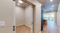 This single-floor space presents a clean, contemporary design throughout, unified by light hardwood flooring and neutral warm-toned walls. The kitchen is a functional highlight, featuring a two-tone cabinetry scheme that pairs charcoal-gray lower cabinets with natural wood-finish upper cabinets. A full suite of stainless steel appliances includes a refrigerator, electric range, over-the-range microwave, and dishwasher, complemented by gray quartz-style countertops and a tile backsplash. Recessed lighting illuminates the space efficiently. The kitchen opens toward a living area with access to a balcony, allowing natural light to flow through. The bedroom is a calm, simply finished room with a window overlooking an adjacent building exterior and neutral walls ideal for customization. The bathroom features a dark espresso-toned vanity with a gray countertop, undermount sink, and a large mirror framed above, paired with chrome fixtures and light hardwood-style flooring. A dedicated storage or utility room, accessible via black-hardware barn-style sliding doors, provides additional functional space and houses in-unit laundry. The overall layout is efficient and well-organized.