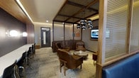 This single-floor lounge space showcases a thoughtfully curated interior design that blends industrial and contemporary aesthetics. The ceiling is a standout architectural feature, incorporating exposed metal mesh panels framed by dark wood beams, complemented by warm cove lighting that runs along the perimeter, creating an inviting ambient glow throughout the space.

The seating area is anchored by a grouping of rich cognac-brown leather armchairs arranged around a low, round wooden coffee table, providing a comfortable and stylish gathering zone. A cluster of matte black spherical pendant lights hangs above, serving as a focal point while delivering targeted illumination. A wall-mounted flat-screen television is positioned nearby, adding a functional media element to the lounge.

Decorative laser-cut wooden or plaster screens on the column and partition add an ornamental detail that contrasts beautifully with the industrial ceiling elements. Wood-paneled walls and partitions contribute warmth and continuity throughout. The flooring features a patterned, multi-toned carpet that adds texture underfoot. A darker private room or secondary space with a glass-paneled door is visible toward the rear, offering additional functionality within the overall layout.