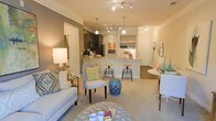 This single-floor residence showcases an open-concept layout that integrates the living, dining, and kitchen areas into one cohesive, flowing space. The living area features plush wall-to-wall carpeting in a neutral tone, providing a soft foundation for the carefully curated furnishings, including a light-upholstered sofa adorned with patterned accent pillows and a rounded ottoman serving as a central focal point.

The kitchen is positioned at the rear of the open layout and features dark wood cabinetry contrasted by a light-toned peninsula with bar-height stools, ideal for casual dining or entertaining. Stainless steel appliances add a modern touch, while pendant lighting above the peninsula and a track lighting system overhead provide layered illumination throughout the space.

Built-in open shelving adjacent to the kitchen offers display and storage opportunities, decorated with tasteful accents. A defined dining area sits to one side, featuring a compact white table. The warm, neutral color palette, combined with soft ambient lighting and cohesive décor, creates an inviting and functional atmosphere throughout the entire space.