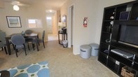 This two-floor residential space in the United States offers a practical and comfortable layout designed for everyday living. The main level features an open-concept living and dining area finished with wall-to-wall carpeting that adds warmth underfoot. The dining area accommodates a table and chairs, making it well-suited for shared meals or casual gatherings. A decorative area rug adds a touch of color to the living space, which also includes a bookshelf unit providing convenient storage and display options. The entryway is bright and welcoming, featuring a paneled front door flanked by a sidelight window that allows natural light to filter into the interior. A small console table with a lamp is tucked neatly near the entry corridor, creating a functional transitional space. Closet storage is accessible from the hallway, offering ample organization options. The neutral, light-toned walls throughout contribute to a clean and airy atmosphere. The space spans two floors, suggesting additional living quarters, bedrooms, or utility areas on the upper level. Overall, the layout is straightforward and efficiently organized, with a calm, neutral palette that provides a versatile backdrop suitable for a variety of furnishing styles and functional needs.