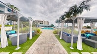 This single-level outdoor space is designed with a resort-inspired aesthetic, centered around a rectangular swimming pool that serves as the focal point of the area. A wide stone-paved walkway leads directly to the pool, flanked on either side by well-maintained artificial turf that adds a clean, vibrant green contrast to the neutral hardscape. Tall palm trees are positioned throughout the space, reinforcing a tropical ambiance and providing natural vertical interest against the open sky.

On both sides of the central pathway, modern pergola-style cabana structures are installed, each draped with flowing white curtains that can be drawn for privacy or shade. Beneath these cabanas, stylish gray wicker lounge furniture with blue and teal accent cushions offers comfortable seating areas ideal for relaxation or social gatherings. The color palette of the space — combining white, gray, teal, and green — creates a cohesive coastal aesthetic throughout.

A light-colored building with teal accents is visible at the far end of the pool area, housing what appears to be a pool house or amenity facility. The overall design of the space prioritizes open-air comfort, stylish outdoor living, and a welcoming, leisure-focused environment.