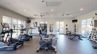 This single-floor fitness center offers a spacious, open layout designed to accommodate a full range of workout routines. The interior is finished with neutral gray walls and durable rubber flooring, creating a clean, functional environment suited for daily exercise. Recessed lighting and multiple ceiling fans are distributed throughout the space, ensuring consistent illumination and airflow during workouts.

The cardio section is well-appointed with True-brand treadmills, elliptical trainers, upright and recumbent stationary bikes, and a rowing machine, offering users a wide variety of low-impact and aerobic exercise options. The strength training area features a multi-station cable machine, a dumbbell rack with a full weight range, a flat workout bench, medicine balls, and stability/exercise balls stored on a dedicated rack.

Large floor-to-ceiling windows and glass French doors line multiple walls, flooding the space with natural light and providing open views of the surrounding outdoor greenery. Full-length mirrors are mounted along one wall, enhancing the sense of space and allowing users to monitor their form. Wall-mounted televisions are positioned throughout the room for entertainment during workouts. The overall ambiance is bright, airy, and well-maintained, offering a comprehensive and inviting fitness experience.