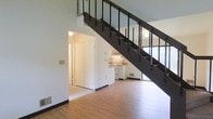 This two-story space is defined by its open, airy design and practical use of vertical space. The main level features light-toned hardwood flooring that extends throughout, creating a warm and cohesive foundation. A bold, dark metal staircase serves as a central architectural element, its open railing design allowing light to pass freely through the space and reinforcing a sense of openness. The high ceilings created by the two-story layout amplify the spacious feel of the main level.

Tucked neatly beneath the staircase is a compact kitchen, efficiently utilizing what might otherwise be unused space. The kitchen features white cabinetry, a range, and under-cabinet lighting, giving it a clean and functional appearance. Adjacent to the kitchen, a closeted area provides additional storage, while a doorway leads to a separate room, adding to the overall functionality of the main floor. Natural light enters through a window visible near the base of the staircase, brightening the lower level. The overall design balances clean, white walls with contrasting dark metalwork, resulting in a contemporary and well-organized living environment.