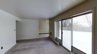 This single-floor space features an open-concept living area finished with neutral-toned walls and smooth white ceilings that lend a clean, understated aesthetic. The room is carpeted in a muted gray tone, contributing to a quiet, low-maintenance interior. A standout feature is the large sliding glass door unit along one wall, which fills the space with natural light and provides direct access to an outdoor patio area. Views through the glass reveal a brick exterior wall and a wintery outdoor setting, suggesting a private, enclosed patio space. A baseboard heating unit runs along the exterior wall beneath the sliding doors, offering an efficient heat source. A wall-mounted heating or ventilation unit is also visible, adding to the climate control options within the space. An open doorway leads into an adjacent room, suggesting a connected layout that allows for easy flow between areas. A wall-mounted light fixture provides additional ambient lighting. Overall, the space presents a functional and straightforward layout with practical finishes and good access to natural light.