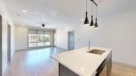 This well-appointed 2-bedroom, 2-bathroom unit offers a thoughtfully designed single-level layout with modern finishes throughout. The open-concept kitchen and living area serves as the heart of the home, anchored by a striking two-tone island with a white quartz countertop and a bold blue base, complemented by grey shaker-style cabinetry, a decorative patterned tile backsplash, and stainless steel appliances. A trio of sleek black pendant lights hangs above the island, adding a contemporary accent. The living area extends generously toward a large bank of windows dressed with roller shades, inviting abundant natural light and outdoor views, while a ceiling fan adds comfort overhead. Light wood-tone flooring flows throughout the main living spaces, contrasting with plush carpeting in the bedrooms. The primary bedroom features a ceiling fan, direct access to its en-suite bathroom, and a spacious walk-in closet outfitted with built-in shelving and hanging rods. The bathrooms are finished with grey cabinetry, white quartz vanities, and large-format tile surrounding the bathtub. A dedicated laundry room with a full-size washer and dryer rounds out this functional and stylish home.