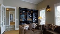 This single-floor space exudes a refined, classic ambiance centered around a thoughtfully designed living area. The room's most commanding feature is a large, dark-finished built-in bookcase that spans an entire wall, offering generous shelving space displayed with books, decorative objects, and curated accessories that lend the room an intellectual warmth. Crown molding along the ceiling adds an architectural elegance, complementing the neutral, earthy wall tones that create a calm and inviting atmosphere throughout. Natural light filters in through a window fitted with slatted blinds, casting a soft glow that highlights the rich textures and tones of the space. Comfortable tufted sofas upholstered in warm brown fabric are arranged invitingly, accented by geometric-patterned throw pillows in muted blue, gold, and cream tones. A slender side table topped with a warm-toned table lamp adds both function and a cozy ambiance to the seating arrangement. The flooring features a light-toned carpet that ties the room together harmoniously. An adjacent hallway, visible through an open doorway, hints at additional rooms beyond, suggesting a thoughtfully connected and well-proportioned overall floor plan. This space blends comfort and classic style seamlessly.