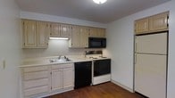 This single-story space includes a well-organized kitchen that serves as a central feature. The kitchen is outfitted with a full suite of appliances, including an electric range with coil burners, an over-the-range microwave, a black dishwasher, and a full-sized refrigerator positioned along the adjacent wall. Light-toned wood cabinetry lines the upper and lower walls, offering generous storage space, while a continuous countertop provides practical workspace. A single-basin sink with a standard faucet is integrated into the counter, positioned beneath the upper cabinets with an under-cabinet light for task lighting. The flooring transitions from dark hardwood in the kitchen area to lighter tiling near the entryway, adding visual contrast. Neutral-toned walls and a textured ceiling contribute to a clean, understated aesthetic throughout. The layout is efficient, with appliances and cabinetry arranged to maximize functionality within the space. Overall, the kitchen presents a practical and neatly maintained environment suitable for everyday use.