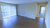 This single-floor space features a spacious, open living area finished with rich, dark hardwood flooring that runs throughout, lending a warm and grounded aesthetic to the interior. The walls are painted in a neutral, light beige tone that reflects natural light and creates a clean, versatile backdrop suitable for a variety of furnishing styles. A large sliding glass door fitted with vertical blinds dominates one wall, allowing abundant natural light to fill the room while providing access to an outdoor area. Glimpses of mature trees are visible through the glass, offering a pleasant natural view. A white baseboard heater runs along the lower portion of the main wall, providing an efficient and unobtrusive heating solution. The layout is open and unobstructed, offering generous usable floor space. A doorway visible on the right side of the room leads to an adjacent area, suggesting a connected, functional floor plan. The overall ambiance is bright, clean, and straightforward, with a neutral palette that makes the space highly adaptable for both personal and professional use.