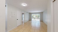 This single-floor space features a cohesive, light-filled design with warm beige ceramic tile flooring running throughout every room, creating a unified and easy-to-maintain aesthetic. The open layout connects the main living area to the kitchen and additional rooms in a straightforward, functional arrangement.

The living area is generously proportioned, with neutral light-gray walls and a flush-mount ceiling fixture providing even illumination. A full-width sliding glass door dominates one wall, fitted with vertical blinds and offering direct views of lush outdoor greenery, allowing abundant natural light to fill the space. A closet with louvered doors is conveniently positioned nearby.

The kitchen is efficiently designed with white cabinetry featuring black hardware, white countertops, a single-basin sink, and stainless steel appliances including a refrigerator and electric range. The compact yet practical layout makes good use of the available space, with a dedicated entry door providing direct exterior access.

The bathroom presents a clean, minimalist aesthetic with fully white square-tiled walls surrounding a tub-shower combination. A white vanity with an integrated sink, black-framed mirror, and black fixtures complete the look, contributing a modern contrast to the all-white palette. Overall, the space maintains a bright, neutral, and well-maintained character throughout.