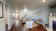 This single-floor unit offers a thoughtfully arranged layout connecting several functional spaces with a cohesive, light aesthetic. The living area sets a welcoming tone with soft blue-grey walls, rich dark hardwood flooring, and a textured ceiling. A contemporary chandelier anchors the space above a comfortable seating arrangement, while decorative wall art and sunburst accents add character. A pass-through window connects the living area directly to the kitchen, enhancing the open feel.

The kitchen features an abundance of white cabinetry with brushed nickel hardware, light stone-look countertops, and a full suite of black appliances including a gas range, built-in microwave, and dishwasher. A double sink sits beneath a mirrored backsplash that reflects natural light into the space.

The bedroom offers a calm retreat with soft carpeting, gentle blue-grey walls, and a textured ceiling, providing a quiet and comfortable atmosphere. The dedicated office space includes carpeted flooring, a window allowing natural light, and ample room for a full work setup.

The bathroom maintains the unit's clean aesthetic, featuring a vanity with light stone countertops, white cabinetry, a large mirror, and a shower-tub combination with a crisp white curtain and hardwood-style flooring.