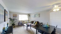 This single-floor residence offers a comfortable and cohesive living environment designed with both functionality and style in mind. The living area serves as the heart of the home, bathed in natural light streaming through a large window that illuminates the soft, neutral-toned carpeting and light-colored walls. The space is thoughtfully arranged to accommodate both relaxation and entertaining, with a plush sofa accented by colorful decorative pillows, a complementary armchair, and a central coffee table. Lush indoor plants add a refreshing touch of greenery, enhancing the welcoming ambiance. Decorative wall art in coastal and abstract styles lends the room a curated, personalized character. A dining area flows naturally from the living space, featuring a dark-toned table and chairs that provide a tasteful contrast to the room's lighter palette. The overall design reflects a clean, open aesthetic with a warm and livable feel, making this a practical yet inviting space suited for everyday living.