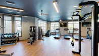Located at 4501 N Garland Ave in Garland, Texas, this fitness center offers a generously sized, single-floor layout designed to accommodate a comprehensive range of workout activities. The space is divided into distinct functional zones, each thoughtfully arranged to maximize usability and flow.

The cardio area features a row of treadmills, stair climbers, and elliptical machines positioned along large windows that flood the space with natural light and provide views of the surrounding outdoors. A bold deep-blue painted ceiling with ceiling fans adds a dynamic visual contrast to the lighter walls and warm wood-style flooring found throughout.

A dedicated cycling studio showcases multiple stationary bikes beneath a tray ceiling with a modern geometric light fixture, complemented by wall-mounted flat-screen TVs for an engaging workout experience. A rack of kettlebells and stacked step platforms provide additional free-weight and functional training options in this area.

The strength and functional training zone is equipped with a multi-functional cable and power rack system, a heavy bag, TRX suspension trainers, and selectorized strength machines. Plyometric boxes offer versatility for high-intensity training. Recessed and linear LED lighting throughout ensures the entire space is well-lit. A built-in decorative shelving unit near the entrance adds a welcoming aesthetic touch, while glass-panel doors connect the various zones seamlessly.