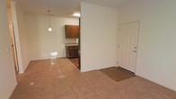 This single-floor space presents a practical and cohesive layout designed for comfortable everyday living. The main living area greets visitors with warm, speckled terrazzo-style flooring that extends throughout the space, lending a clean and timeless quality to the interior. A white paneled entry door with a brushed nickel handle provides a welcoming entry point, transitioning seamlessly into the open living zone.

A partial wall divides the living area from the kitchen, creating a sense of definition while maintaining an open feel between the two spaces. The kitchen is outfitted with rich, medium-toned wood cabinetry offering ample storage, paired with a dark countertop and stainless steel appliances. Hardwood-style flooring in the kitchen contrasts nicely with the living area flooring, subtly delineating the two zones.

A pendant light fixture hangs above, providing focused task lighting that complements the overhead fluorescent fixture in the kitchen. The neutral cream-toned walls throughout create a bright and airy ambiance, reflecting light effectively across the space. Overall, the layout is efficient and well-connected, making it easy to navigate between the entry, living, and kitchen areas while maintaining a cohesive and inviting atmosphere.