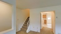 This two-story space is characterized by its clean, understated design and functional layout. The main level features wall-to-wall carpeting in a neutral beige tone, creating a cohesive and understated aesthetic throughout the common areas. A carpeted staircase with a white wooden railing and baluster system connects the two floors, positioned centrally to allow easy access while maintaining an open feel in the surrounding space. The smooth, white-painted walls are kept minimal, offering a blank canvas that maximizes the sense of openness. Electrical outlets and light switch panels are conveniently placed along the walls. Adjacent to the staircase, a doorway leads into a kitchen area, which features warm honey-toned wood cabinetry and what appears to be tile or hard flooring, providing a practical contrast to the carpeted living areas. The kitchen cabinetry includes both upper and lower units, suggesting ample storage. The overall ambiance of the space is neutral and move-in ready, with a straightforward layout that emphasizes practicality. The light, airy color palette throughout contributes to a bright and welcoming atmosphere across both levels.