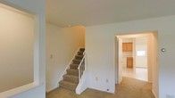 This two-story space is characterized by its clean, understated design and functional layout. The main level features wall-to-wall carpeting in a neutral beige tone, creating a cohesive and understated aesthetic throughout the common areas. A carpeted staircase with a white wooden railing and baluster system connects the two floors, positioned centrally to allow easy access while maintaining an open feel in the surrounding space. The smooth, white-painted walls are kept minimal, offering a blank canvas that maximizes the sense of openness. Electrical outlets and light switch panels are conveniently placed along the walls. Adjacent to the staircase, a doorway leads into a kitchen area, which features warm honey-toned wood cabinetry and what appears to be tile or hard flooring, providing a practical contrast to the carpeted living areas. The kitchen cabinetry includes both upper and lower units, suggesting ample storage. The overall ambiance of the space is neutral and move-in ready, with a straightforward layout that emphasizes practicality. The light, airy color palette throughout contributes to a bright and welcoming atmosphere across both levels.