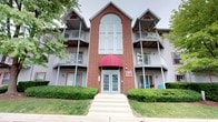 This multi-story residential building presents a classic and well-maintained exterior characterized by a combination of warm red brick and gray siding. The facade is anchored by a striking central brick tower that rises above the roofline, featuring a large arched window that draws natural light into the interior common areas and adds significant architectural interest to the building's profile.

The main entrance is welcoming and clearly defined, framed by a bold burgundy awning and accessed through a set of double glass-panel doors, approached via a wide concrete walkway with steps leading up from the sidewalk. Red accent trim throughout the exterior ties the design elements together cohesively.

Flanking the central tower on both levels are private balconies with metal railings, offering outdoor space for residents. Several balconies are adorned with potted plants and flowers, adding a lived-in, homey character to the building. Ground-floor units feature large windows with decorative red trim, providing ample natural light. Well-maintained shrubs and greenery line the building's perimeter, enhancing its overall curb appeal.