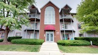 This multi-story residential building presents a classic and well-maintained exterior characterized by a combination of warm red brick and gray siding. The facade is anchored by a striking central brick tower that rises above the roofline, featuring a large arched window that draws natural light into the interior common areas and adds significant architectural interest to the building's profile.

The main entrance is welcoming and clearly defined, framed by a bold burgundy awning and accessed through a set of double glass-panel doors, approached via a wide concrete walkway with steps leading up from the sidewalk. Red accent trim throughout the exterior ties the design elements together cohesively.

Flanking the central tower on both levels are private balconies with metal railings, offering outdoor space for residents. Several balconies are adorned with potted plants and flowers, adding a lived-in, homey character to the building. Ground-floor units feature large windows with decorative red trim, providing ample natural light. Well-maintained shrubs and greenery line the building's perimeter, enhancing its overall curb appeal.