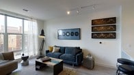 This single-floor space presents a thoughtfully designed living area that balances modern aesthetics with comfortable functionality. The lounge is anchored by a deep blue velvet sofa adorned with mustard yellow and neutral accent pillows, positioned atop a patterned area rug. A sleek, mixed-material coffee table with a glass inset serves as a central focal point.

The walls are kept clean and bright, providing an ideal backdrop for the curated artwork on display, including a framed horizontal print featuring vinyl record motifs and a series of sculptural wall pieces with a metallic, industrial character. Track lighting runs along the ceiling, offering directed illumination throughout the space.

Natural light filters softly through a sheer-curtained window on one side, contributing to the room's airy, open atmosphere. A bold industrial-style floor lamp adds both functional lighting and a design statement. A small side table near the sofa provides practical storage. The light-toned walls and neutral flooring unify the space, creating a cohesive, contemporary ambiance suitable for both relaxing and entertaining.