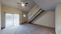 This two-story home offers a thoughtfully designed layout that balances open living spaces with practical functionality. The main level centers around a spacious living area highlighted by soaring vaulted ceilings and a ceiling fan, with a sliding glass door that invites natural light and outdoor views. An octagonal accent window adds a distinctive architectural touch. The kitchen features a wraparound granite-topped peninsula with a built-in sink, warm wood cabinetry, a tile backsplash, and recessed lighting overhead, along with full appliances including a stainless range and black refrigerator. A bedroom and a full bathroom with granite countertops and tile flooring round out the main level. The staircase, fitted with a wood handrail and white spindle balustrade, leads to the upper floor, where a generous loft-style landing is bathed in natural light from a large skylight. The upper level includes additional carpeted bedrooms with windows overlooking the surrounding area, and a second bathroom also featuring granite countertops, a large mirror, and tile flooring. Warm neutral tones, wood trim, and plush carpeting throughout create a cohesive and welcoming atmosphere across both floors.